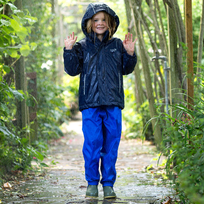 A young child in Originals Waterproof Trousers Blue, stands in a garden in the rain, holding their hands up.